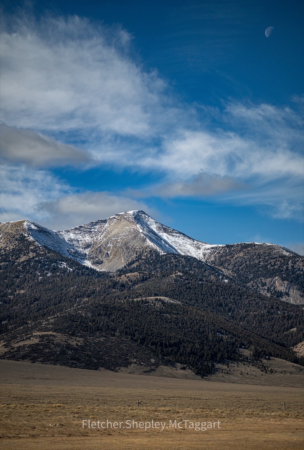 Clouds move over a steep mountain beneath a small moon.