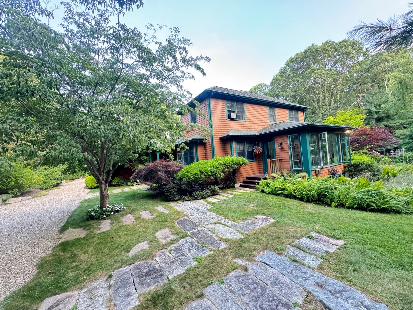 A shingled home with green trim, a glass porch, and stone paths through a mature garden