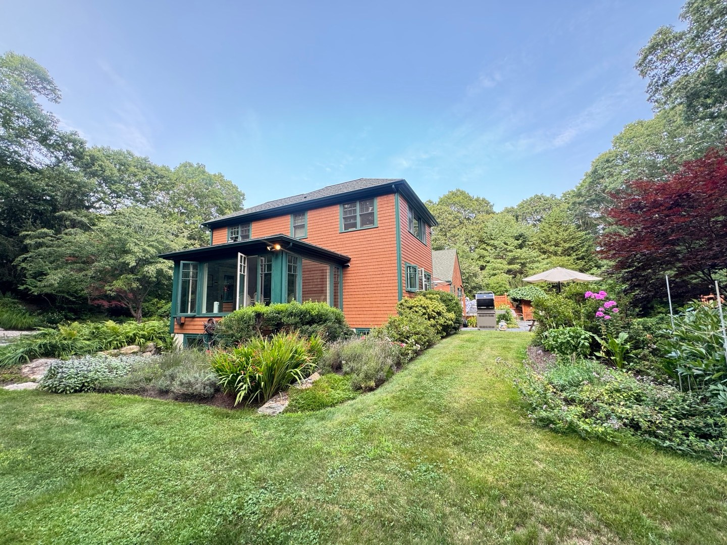 A garden frames the side of a shingled home, with a glass porch and outdoor seating