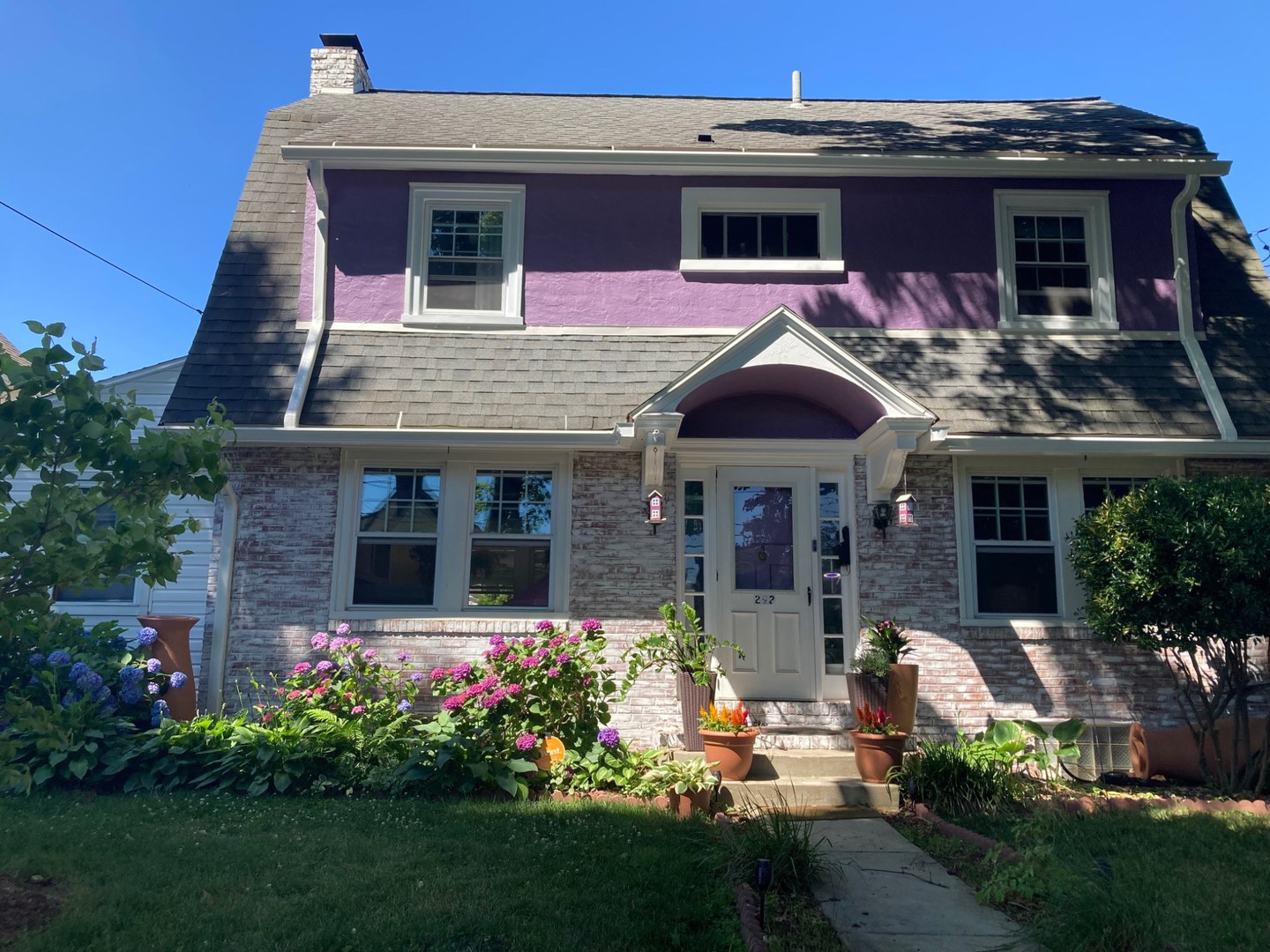 A purple and brick home with a curved porch roof and flowering front garden